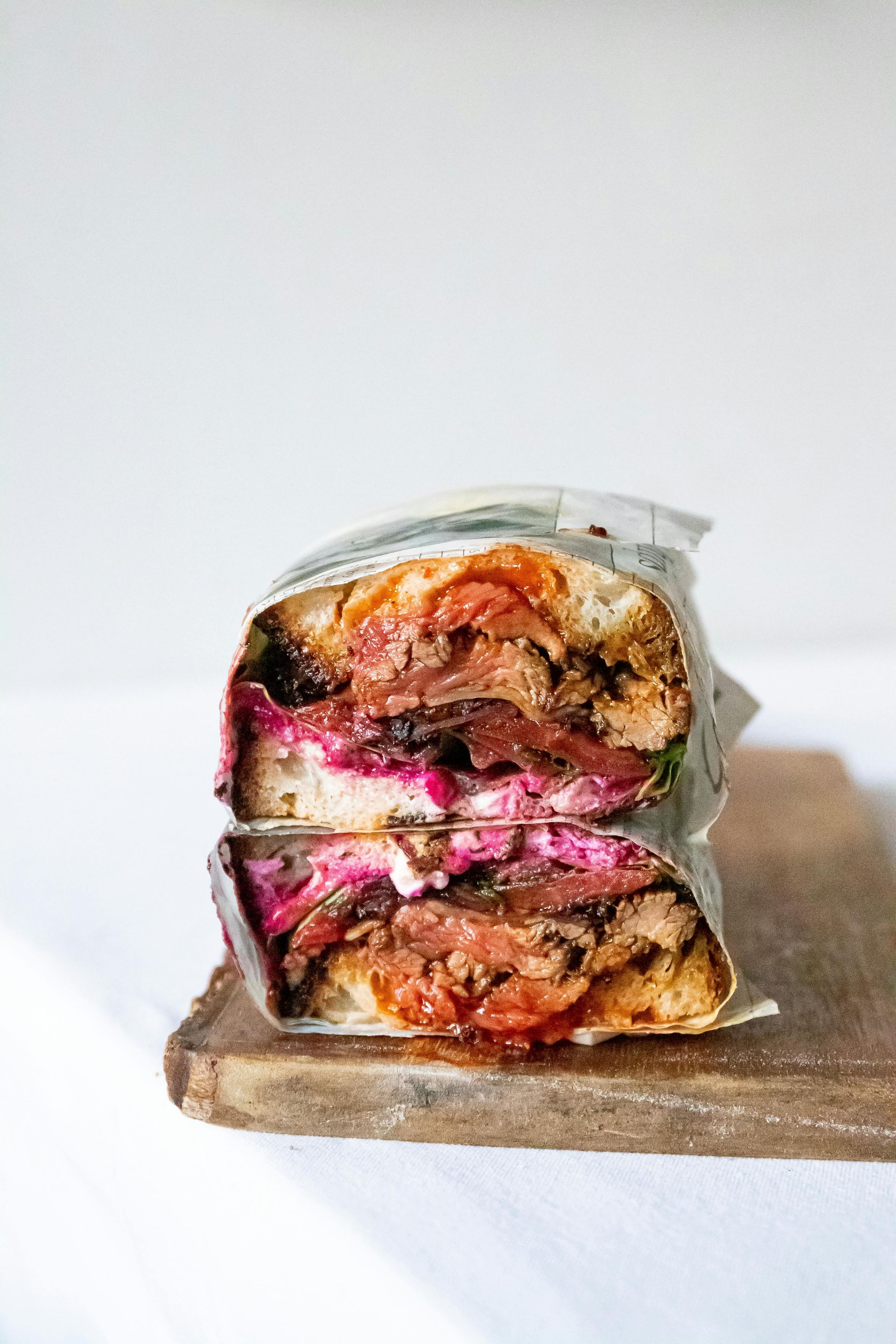 Sandwich cut in half, wrapped in paper, with meat, beets, and sauce on a wooden board.