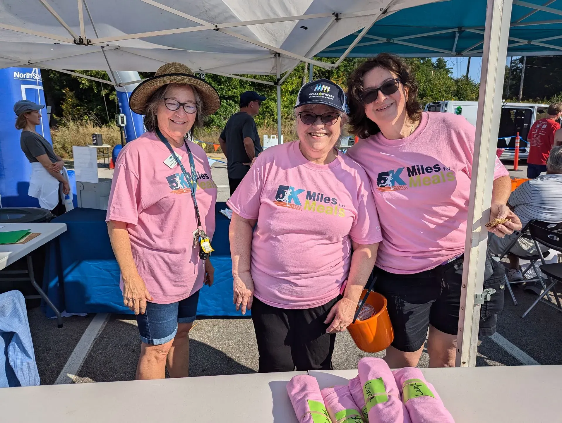Three women in pink shirts smiling at a booth, possibly at an event, with an outdoor setting.