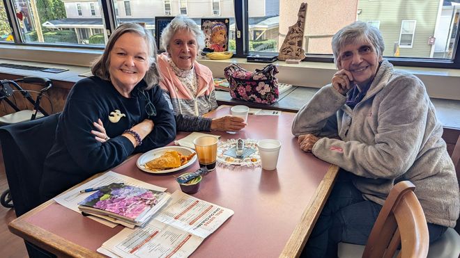 Three people sit at a table in front of a window. Two smile at the camera. Food and drinks on table.