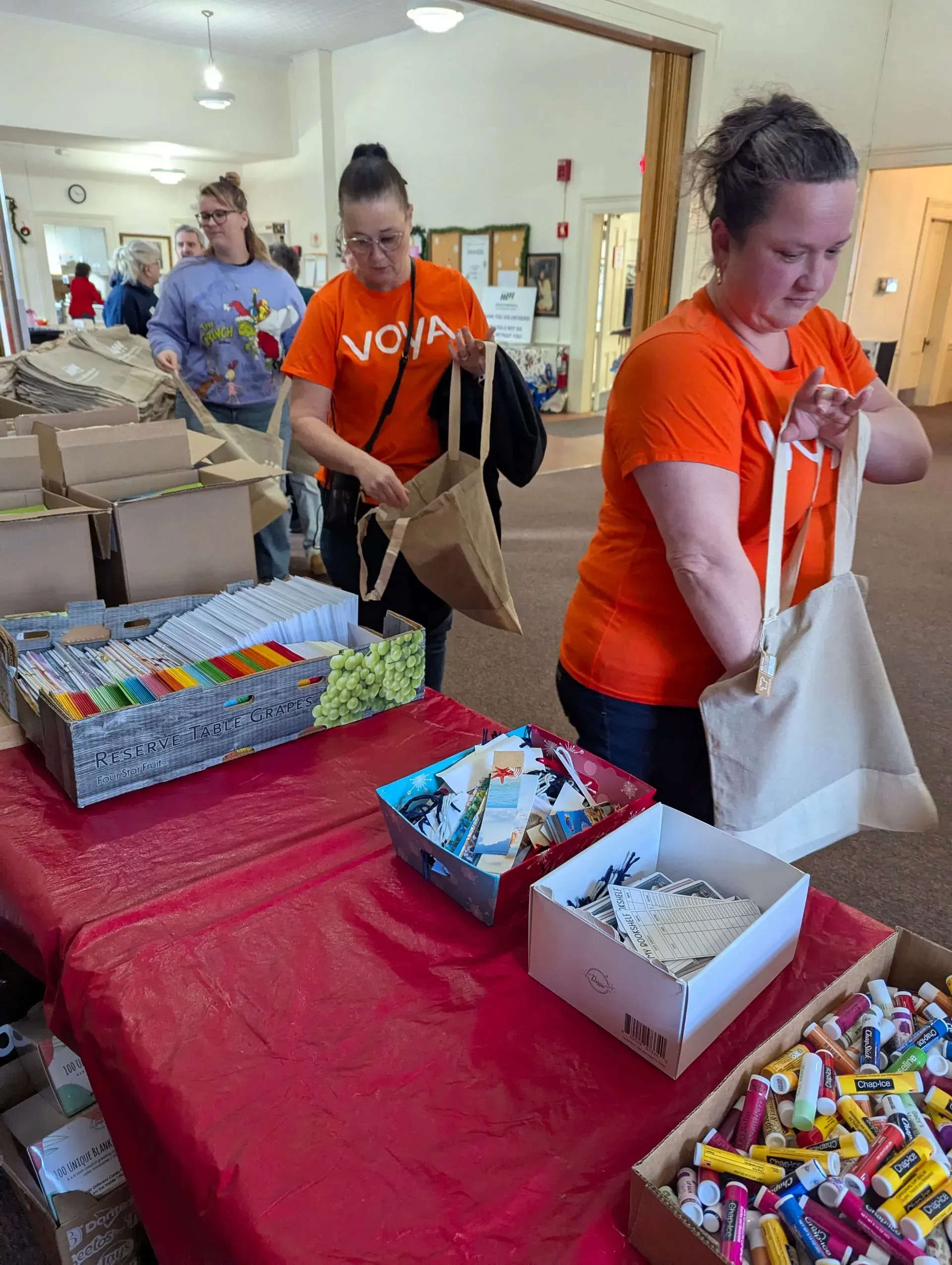 People packing supplies into bags at a table. Volunteers in orange shirts. Boxes and bins of supplies.