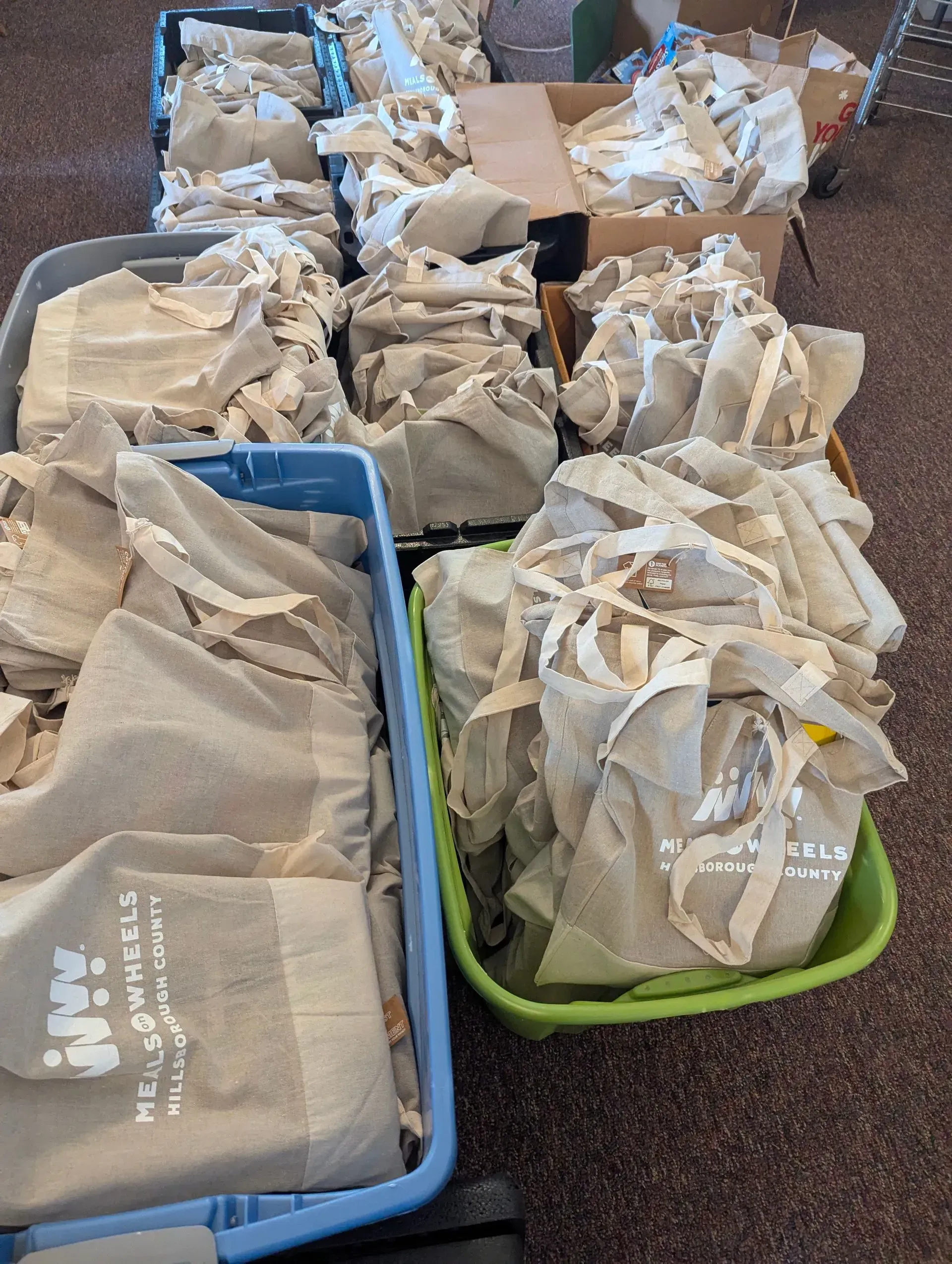 Several beige tote bags, some filled with paper, in blue and green bins and cardboard boxes.
