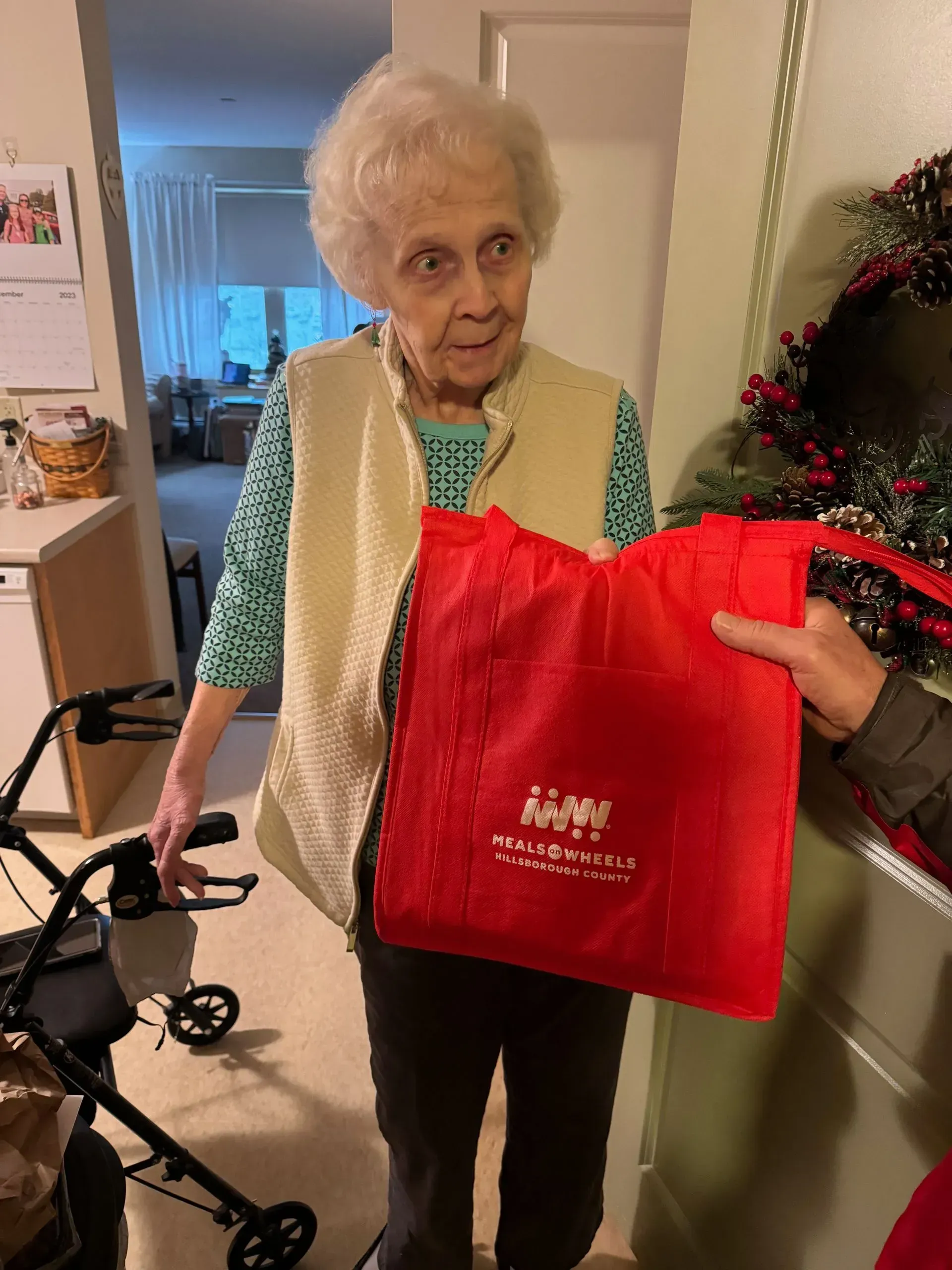 Elderly woman holding a red bag, standing near a walker and decorated door, possibly receiving a gift.