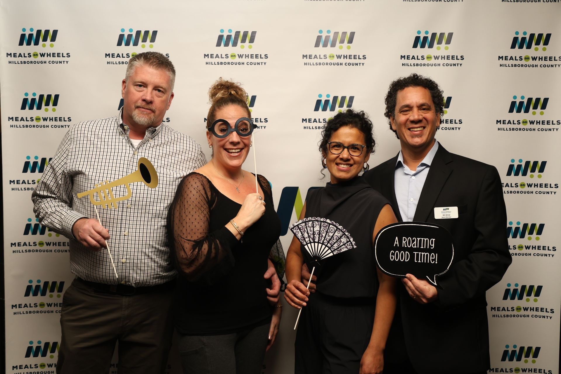 Four people pose at an event with props. Two hold up fake glasses and a horn. A woman holds a fan.