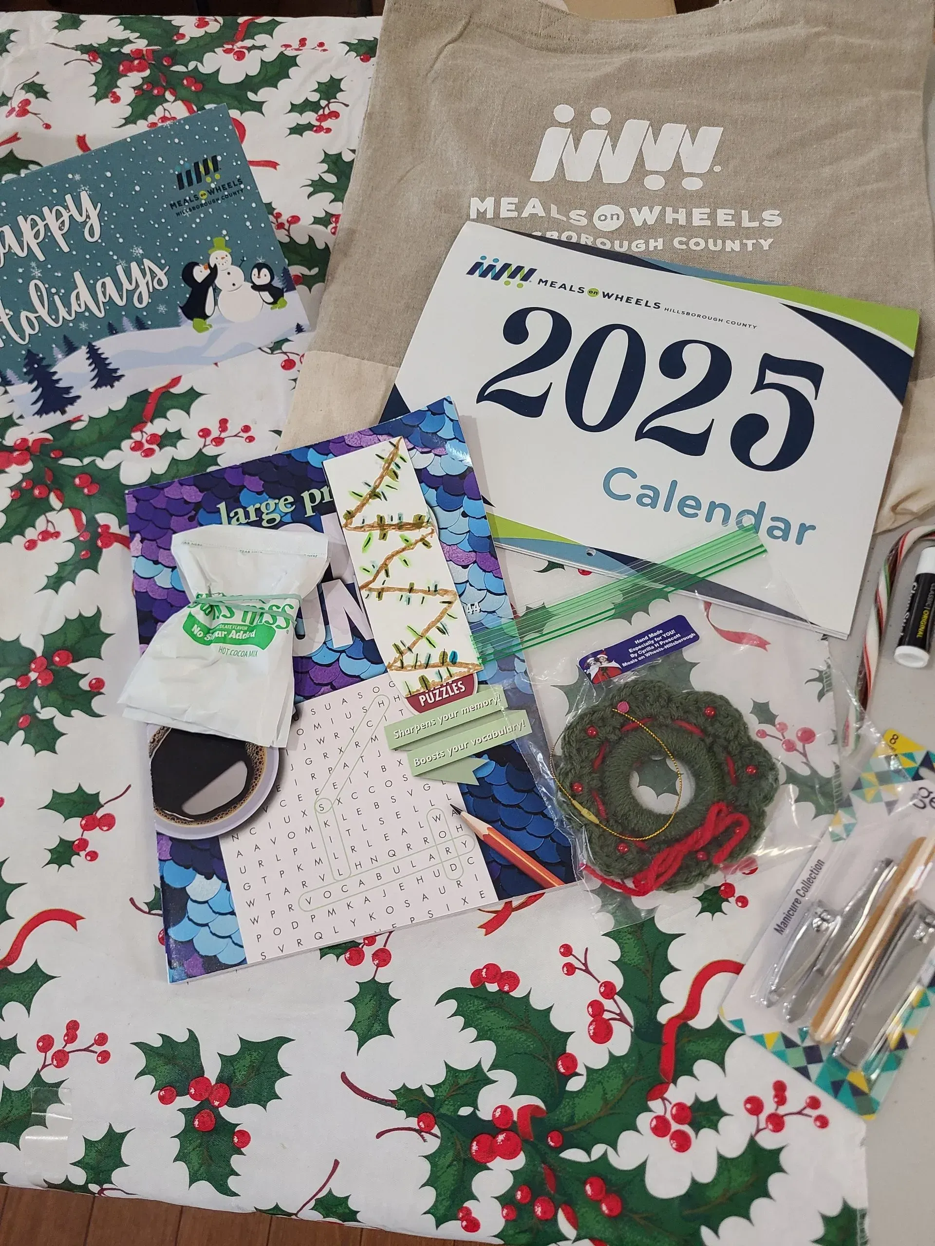 Holiday items on festive tablecloth: card, 2025 calendar, craft supplies, tote bag, and mini wreath.