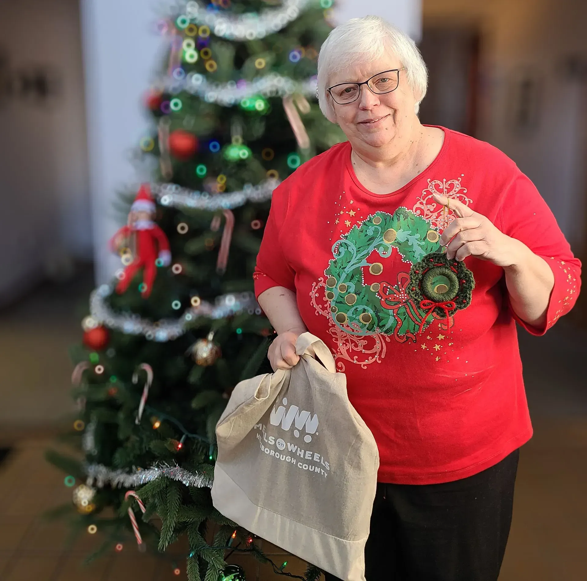 Woman in red shirt with a Christmas wreath decoration holds a tote bag near a decorated tree.
