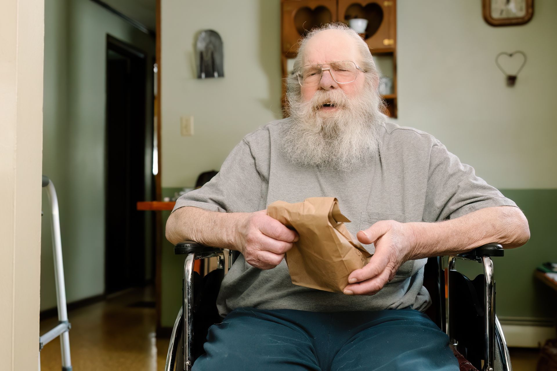 Man with long white beard in a wheelchair holds a brown paper bag, indoors.