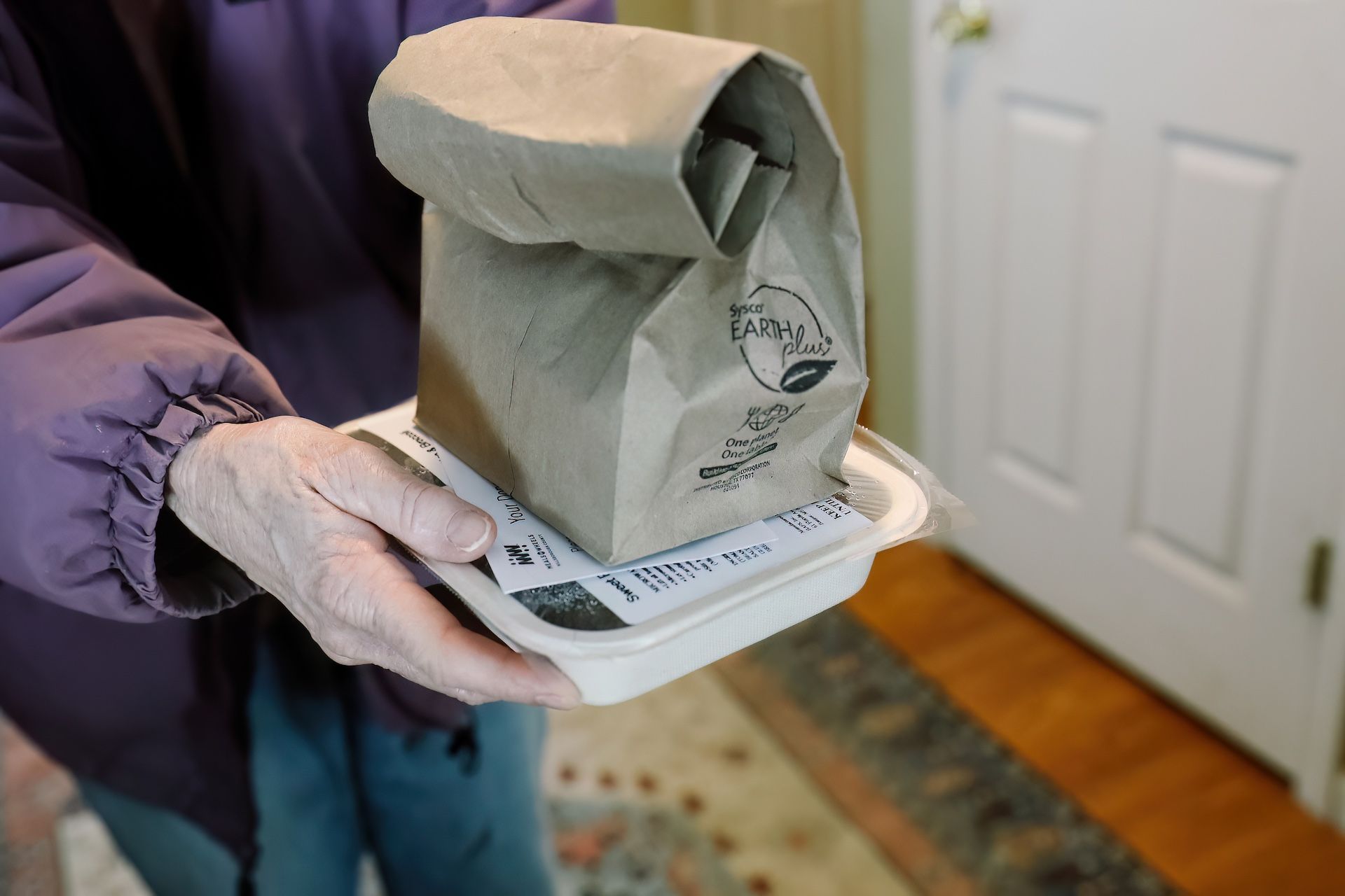 Person holding a brown paper bag with a logo on top of a food container; near a door.