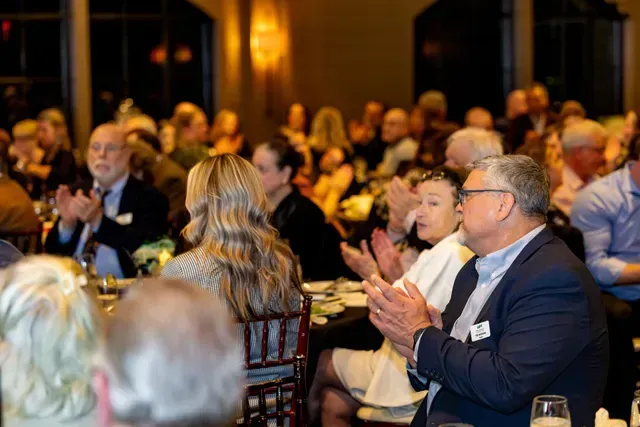 People clapping at a formal event in a dimly lit room, with tables and a crowd.