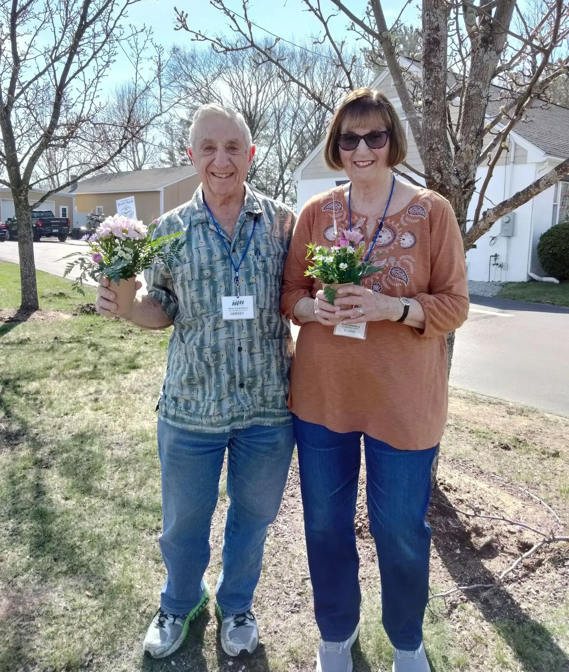 Two people holding potted flowers outside; sunny day.