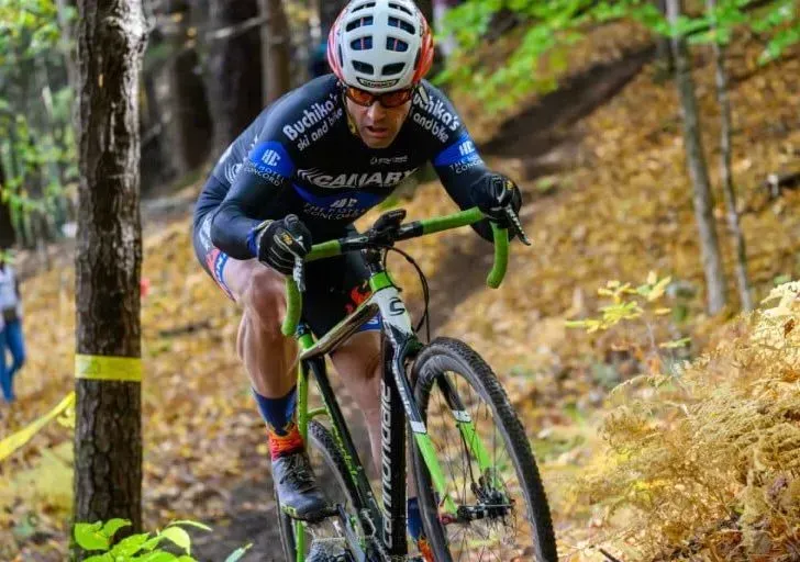 Cyclocross cyclist in black and blue jersey, descending a wooded trail on a green and black bike.