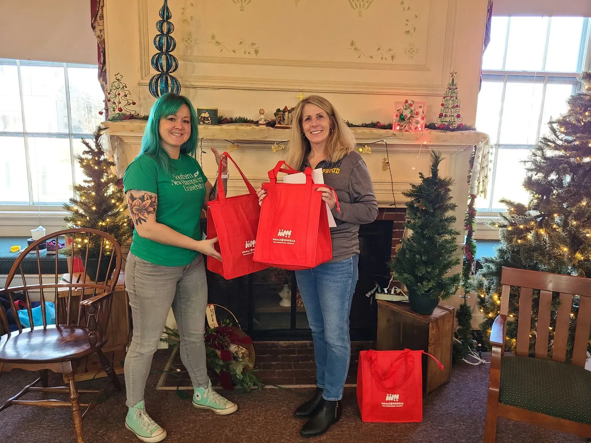 Two women holding red gift bags in front of a decorated fireplace with Christmas trees.