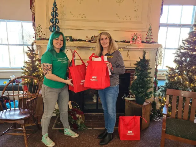 Two women holding red gift bags in front of a decorated fireplace with Christmas trees.