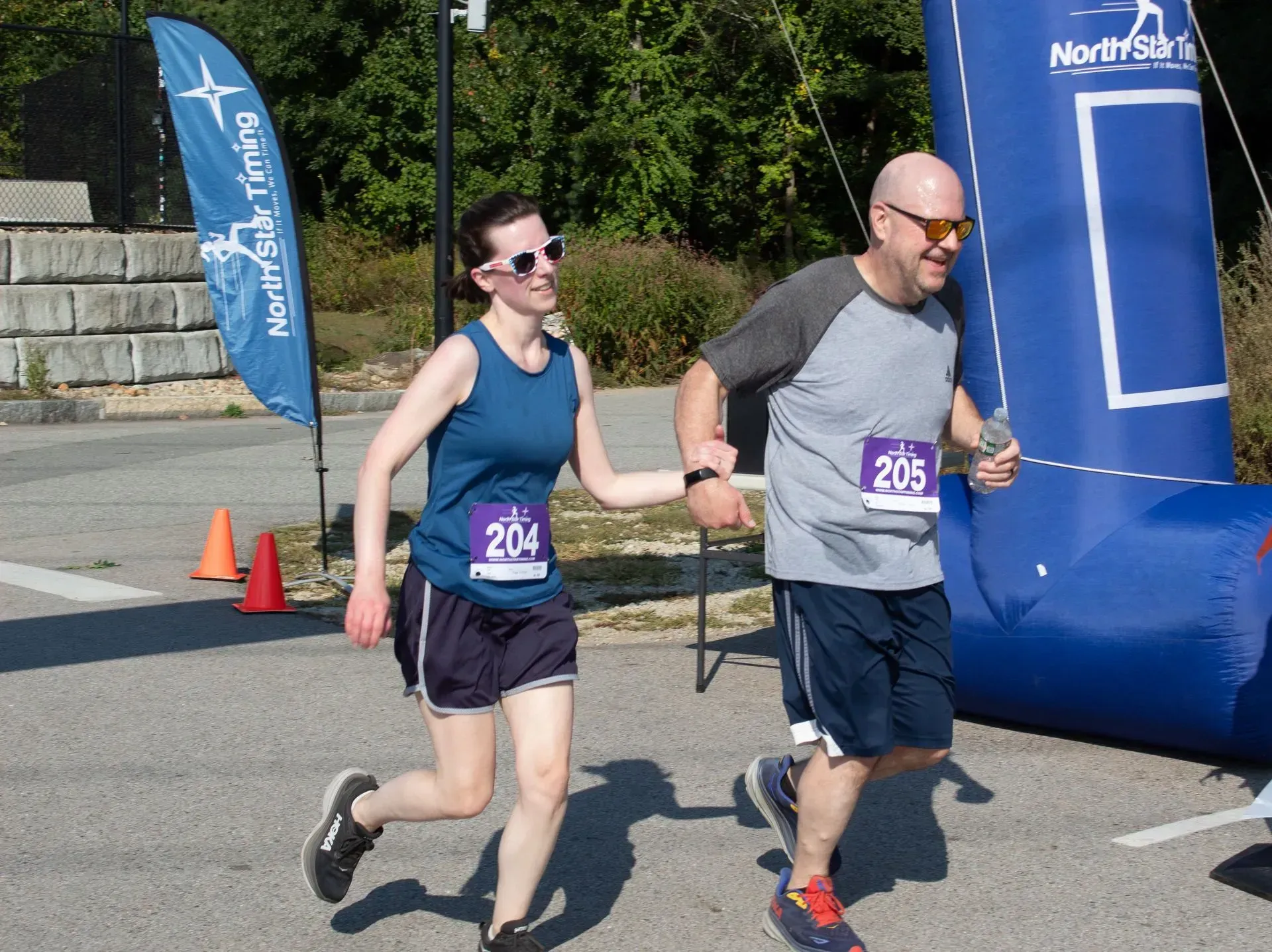 A man and woman running a race, holding hands near the finish line. Outdoors, sunny.
