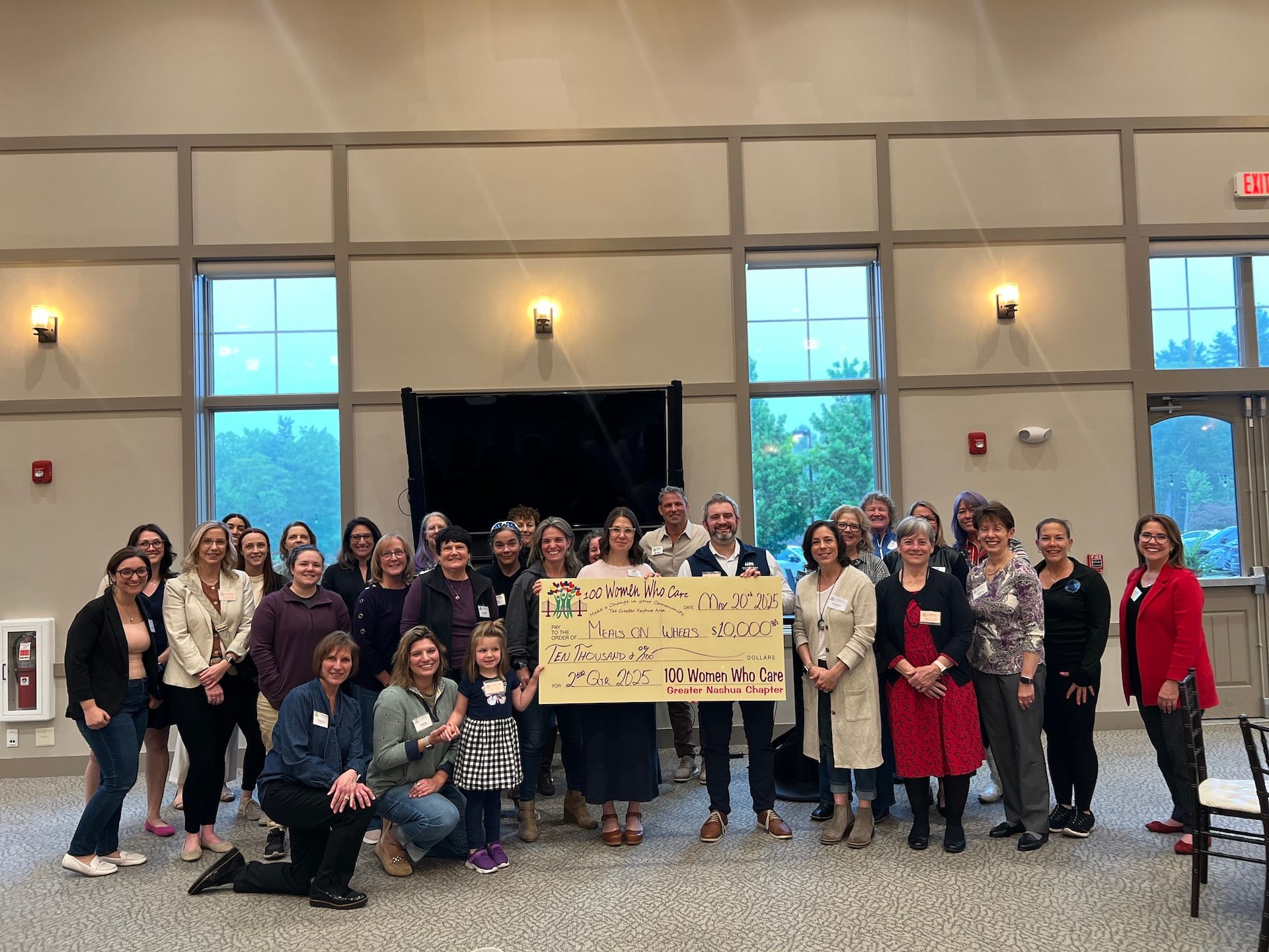 Group of people holding a large check in a bright room with windows.