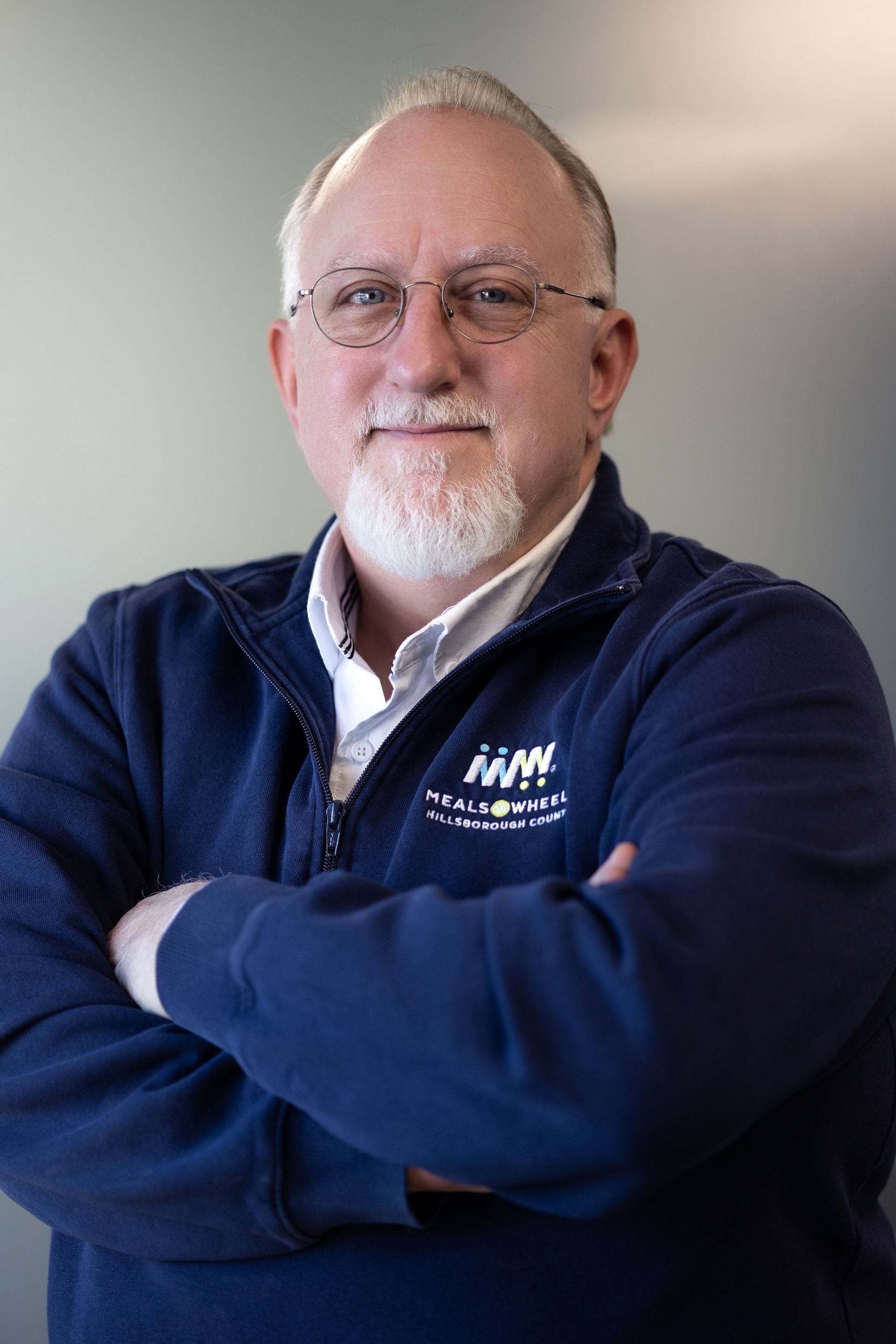 Man with glasses and white beard, arms crossed, wearing a blue sweater, smiling.