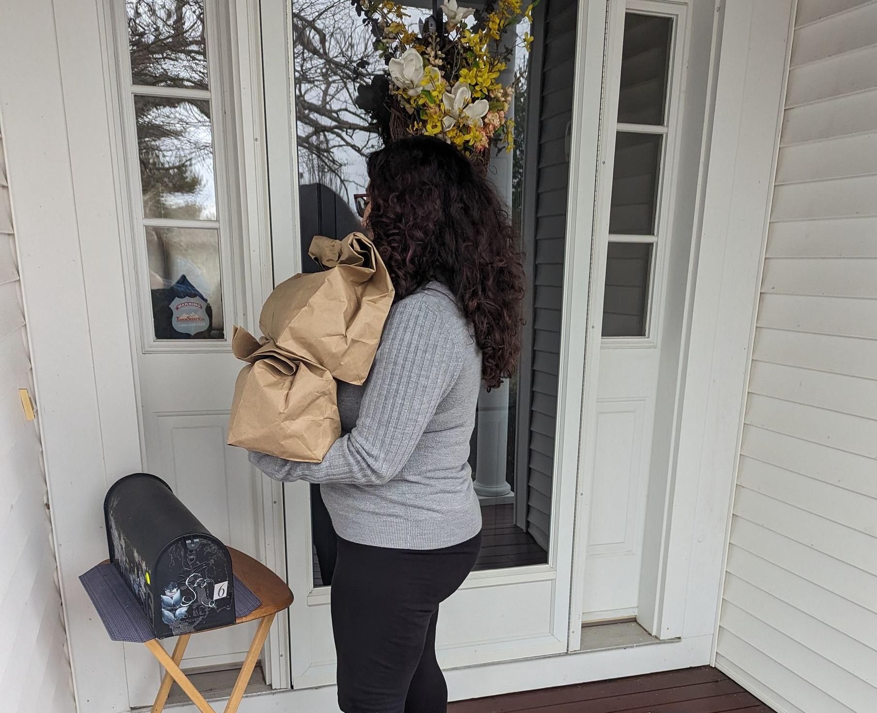 Woman carrying two brown paper bags, standing at a white door with a wreath.