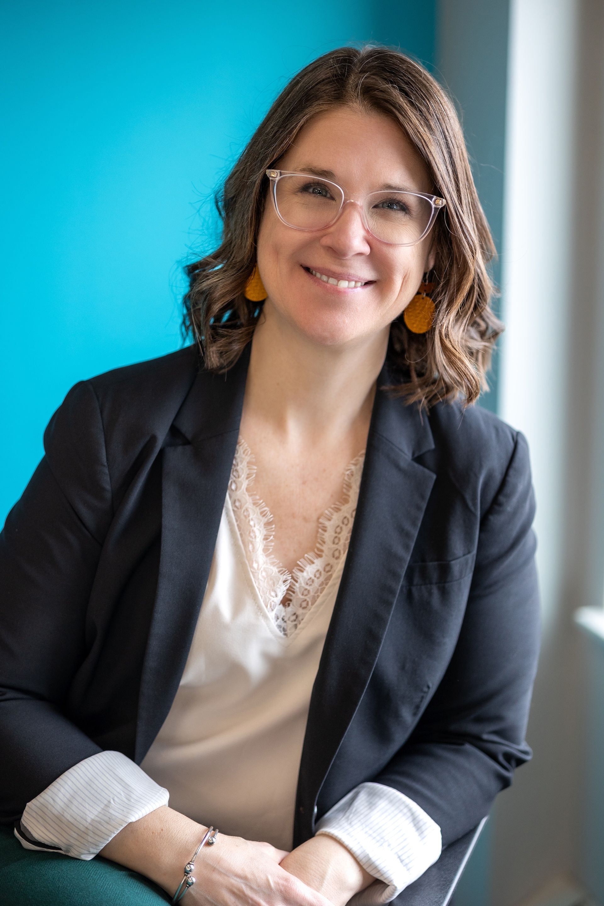 Woman with glasses smiles, wearing a black blazer and white top, seated against a turquoise wall.