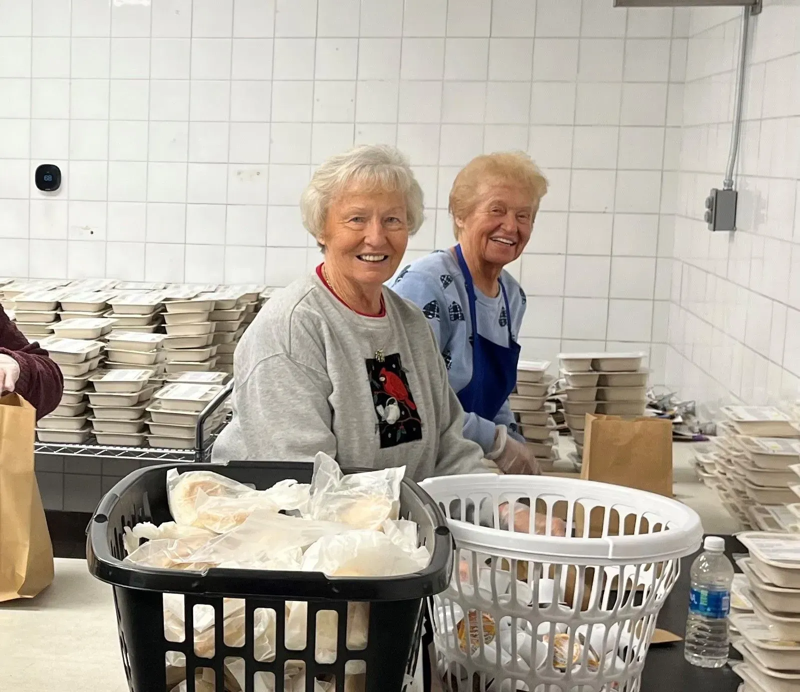 Two smiling women preparing meals in a kitchen, surrounded by stacks of containers and baskets.