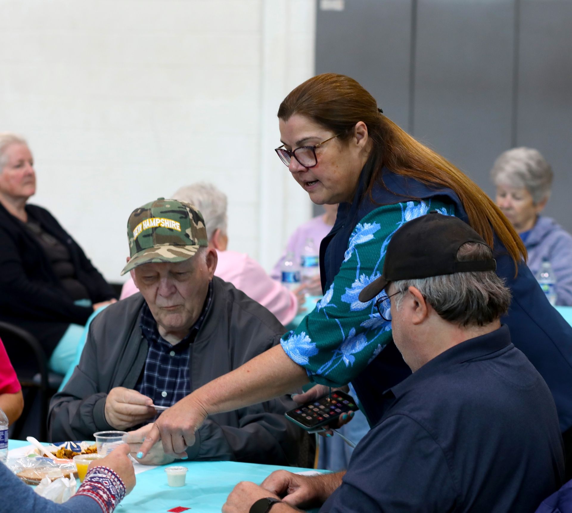 Woman assisting elderly people at a table; some are playing cards, others watching.