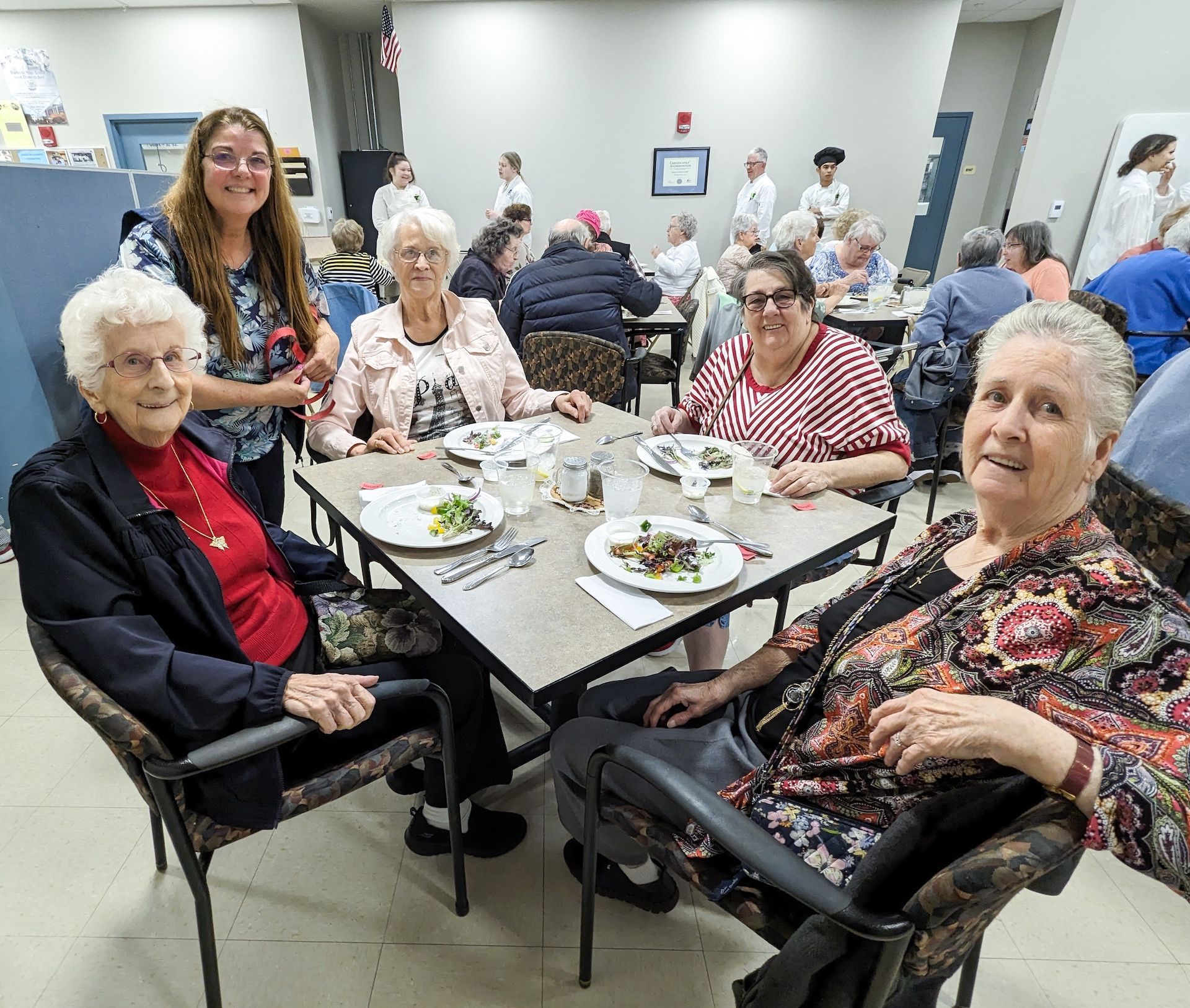 Four women and a staff member smile at a table in a dining hall. Others eat at tables.