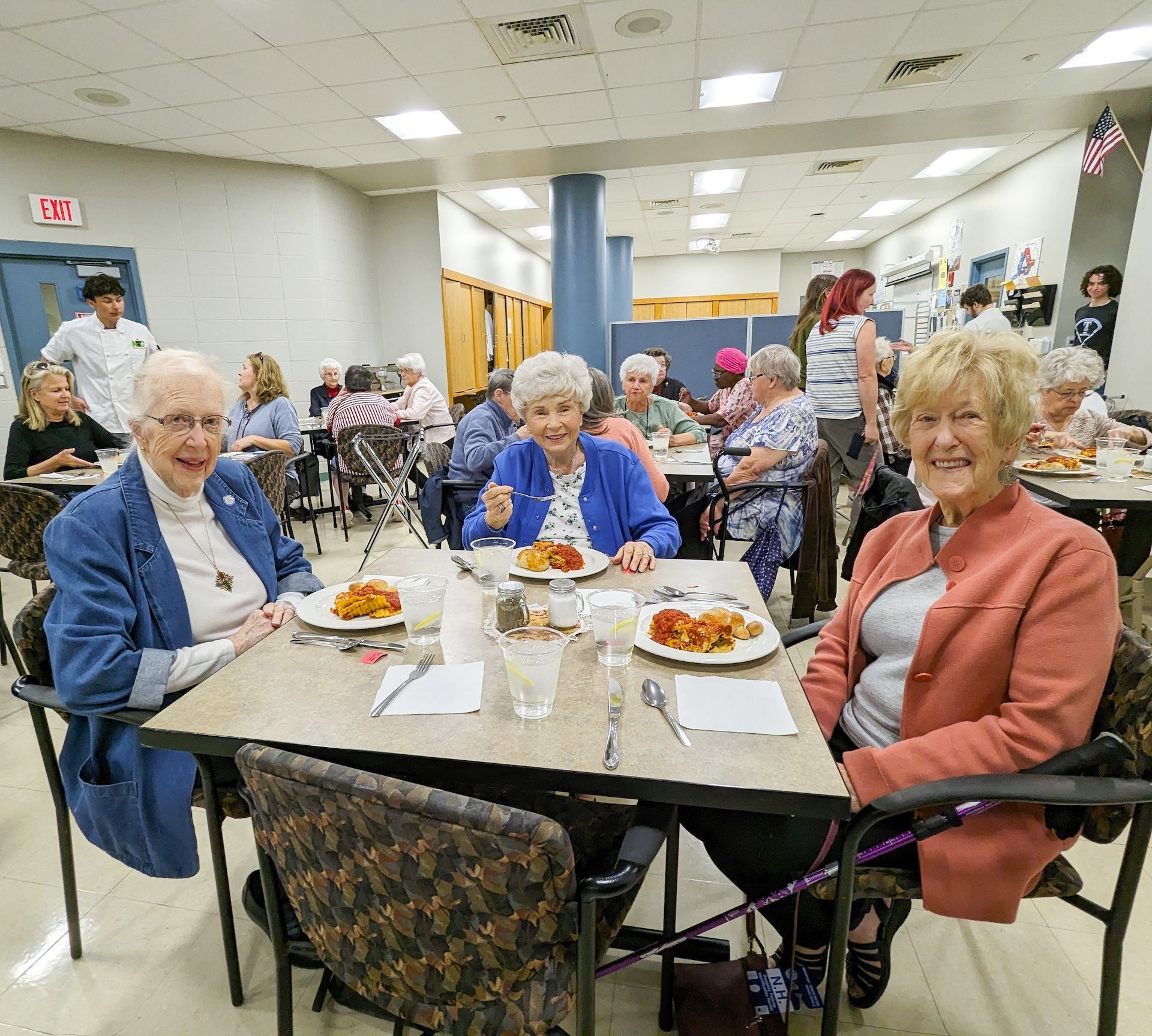Seniors smiling, eating lunch in a bright dining room. People are seated at tables; a server is visible.