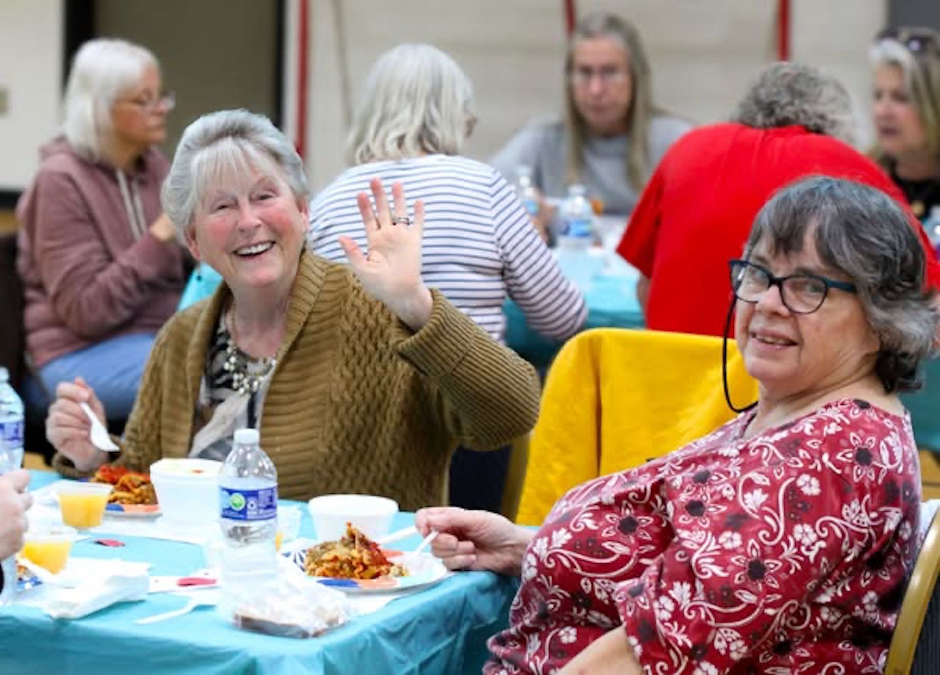 Two older women wave and smile at a table set for a meal; others dine in the background.