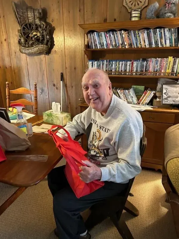 An elderly man with a bright smile, holding a red gift bag, seated at a table indoors.