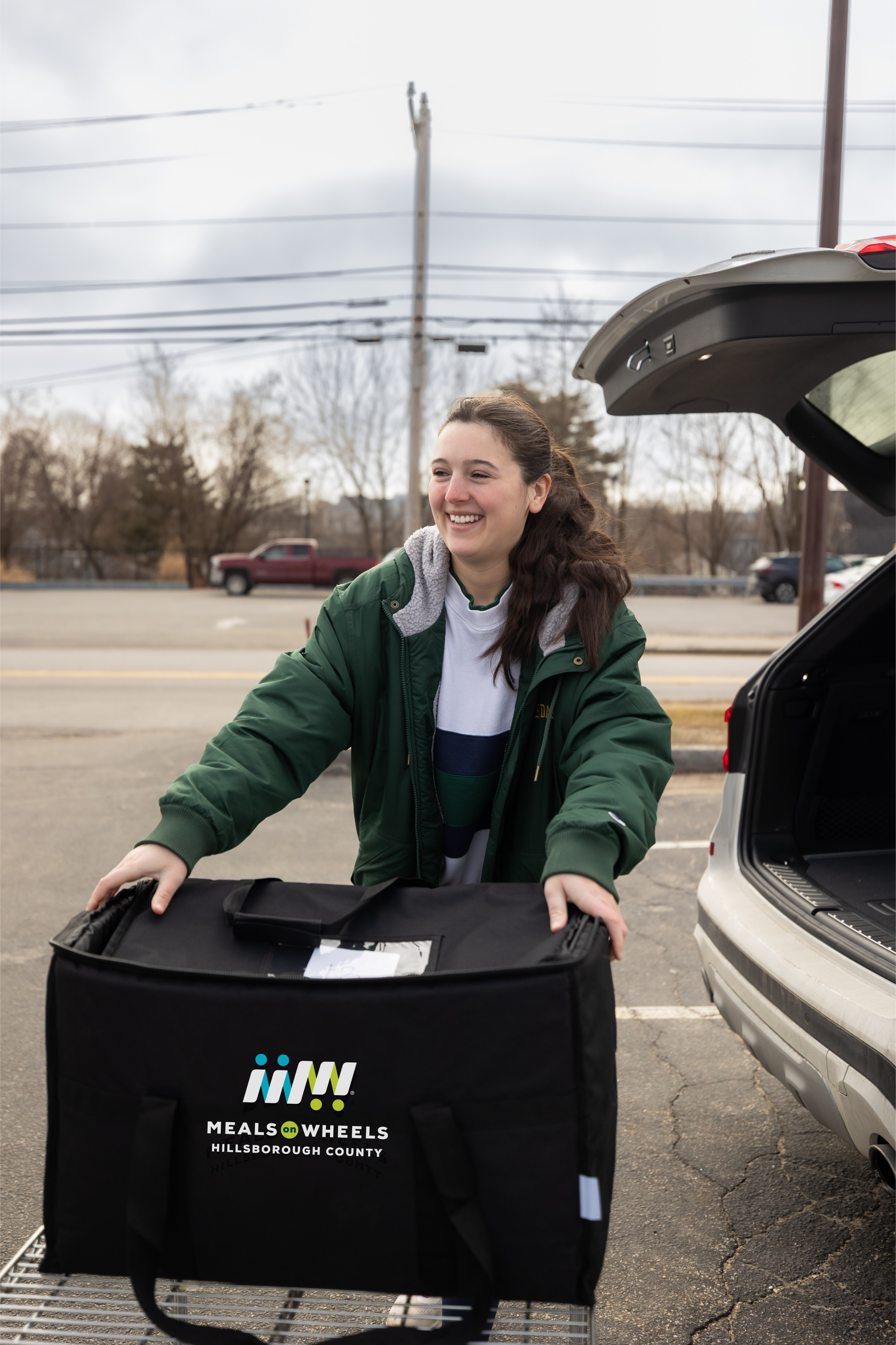Woman smiling, wearing Patriots sweatshirt, holding tablet, in a food pantry setting with volunteers.