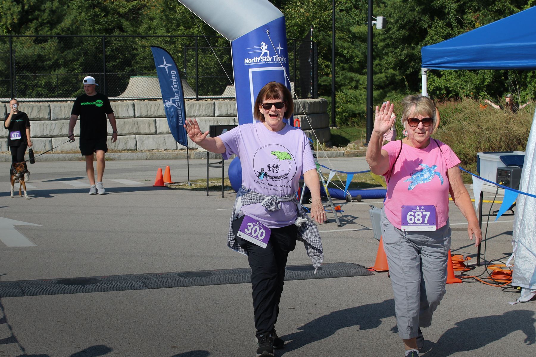 Two women wave, crossing a race finish line. One wears a purple shirt, the other pink. Outdoors, sunny day.