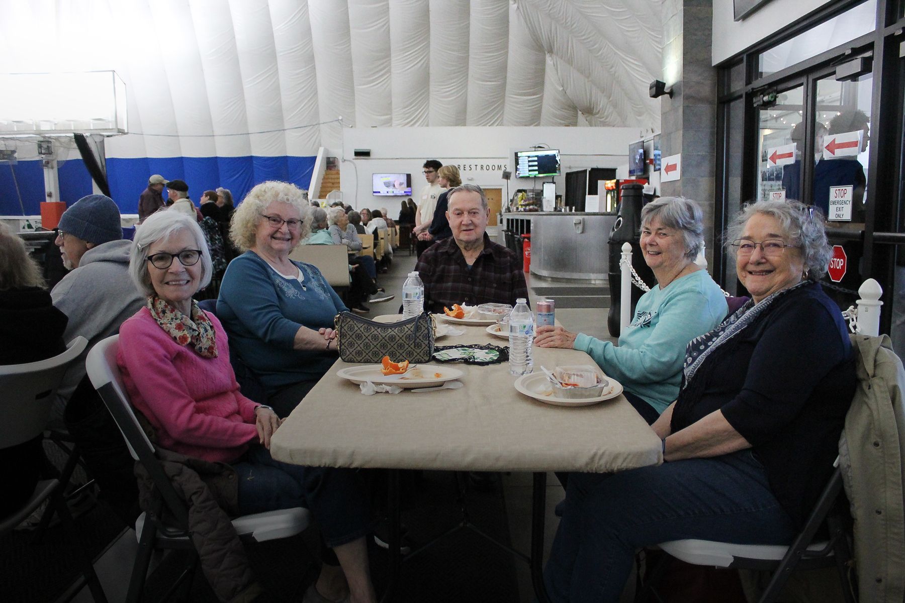Four older adults seated at a table indoors, smiling. Snacks, drinks present. Dome background.