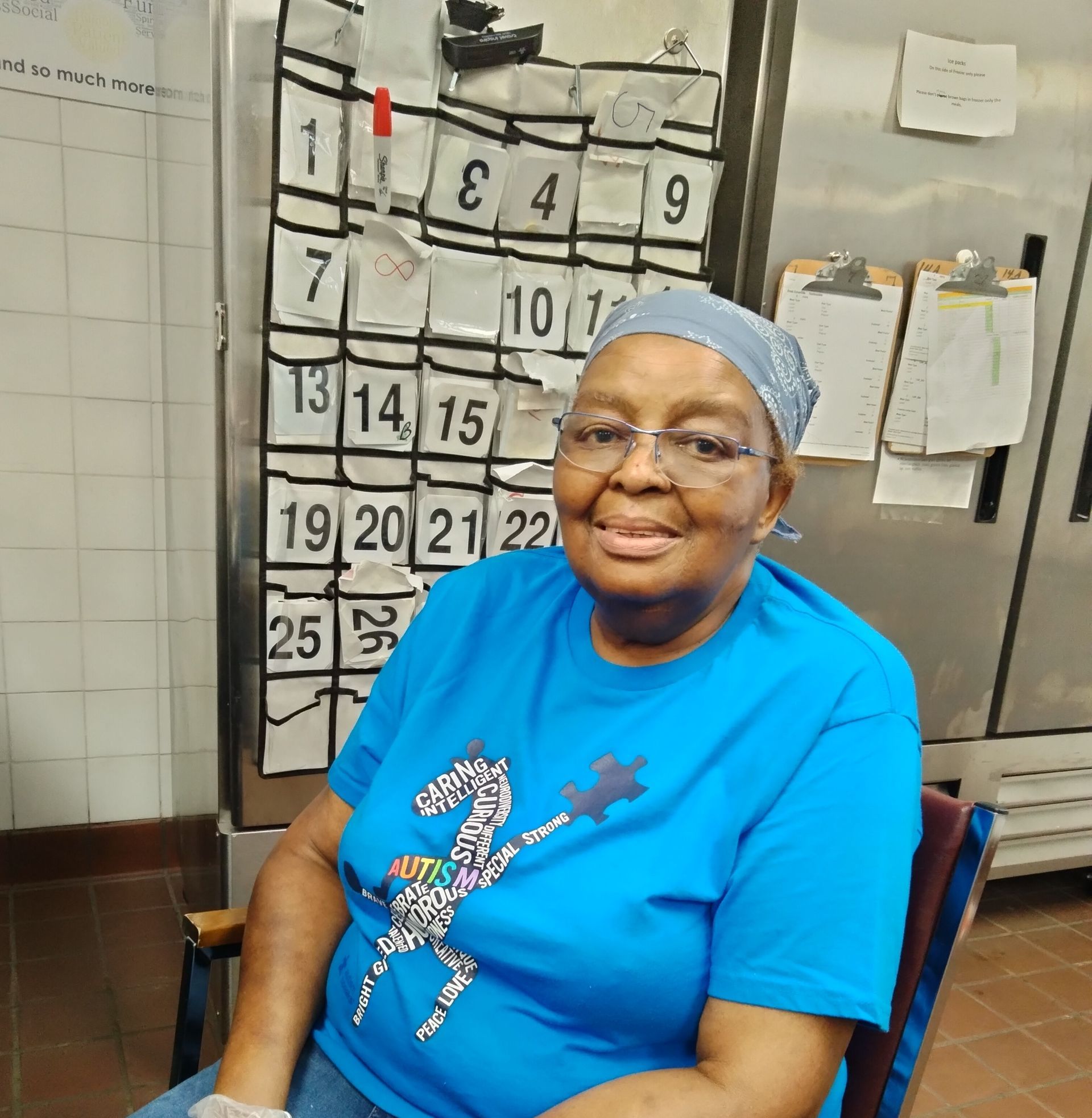 Woman in blue shirt and glasses smiles in a kitchen; a calendar is behind her.