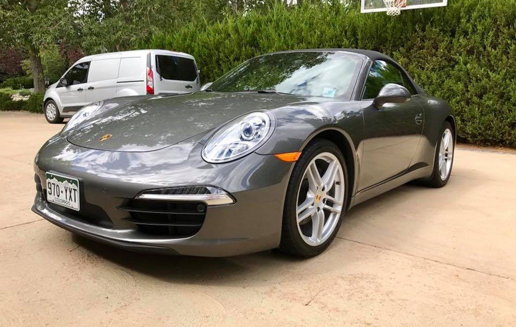 A gray porsche 911 carrera is parked in a driveway next to a basketball hoop.