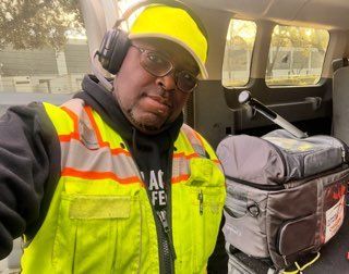 A man wearing a yellow vest and headphones is taking a selfie in the back of a van.