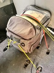 A backpack is sitting on the floor with a yellow strap attached to it.