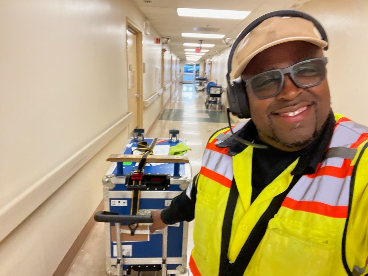A man wearing headphones and a yellow vest is pushing a cart down a hallway