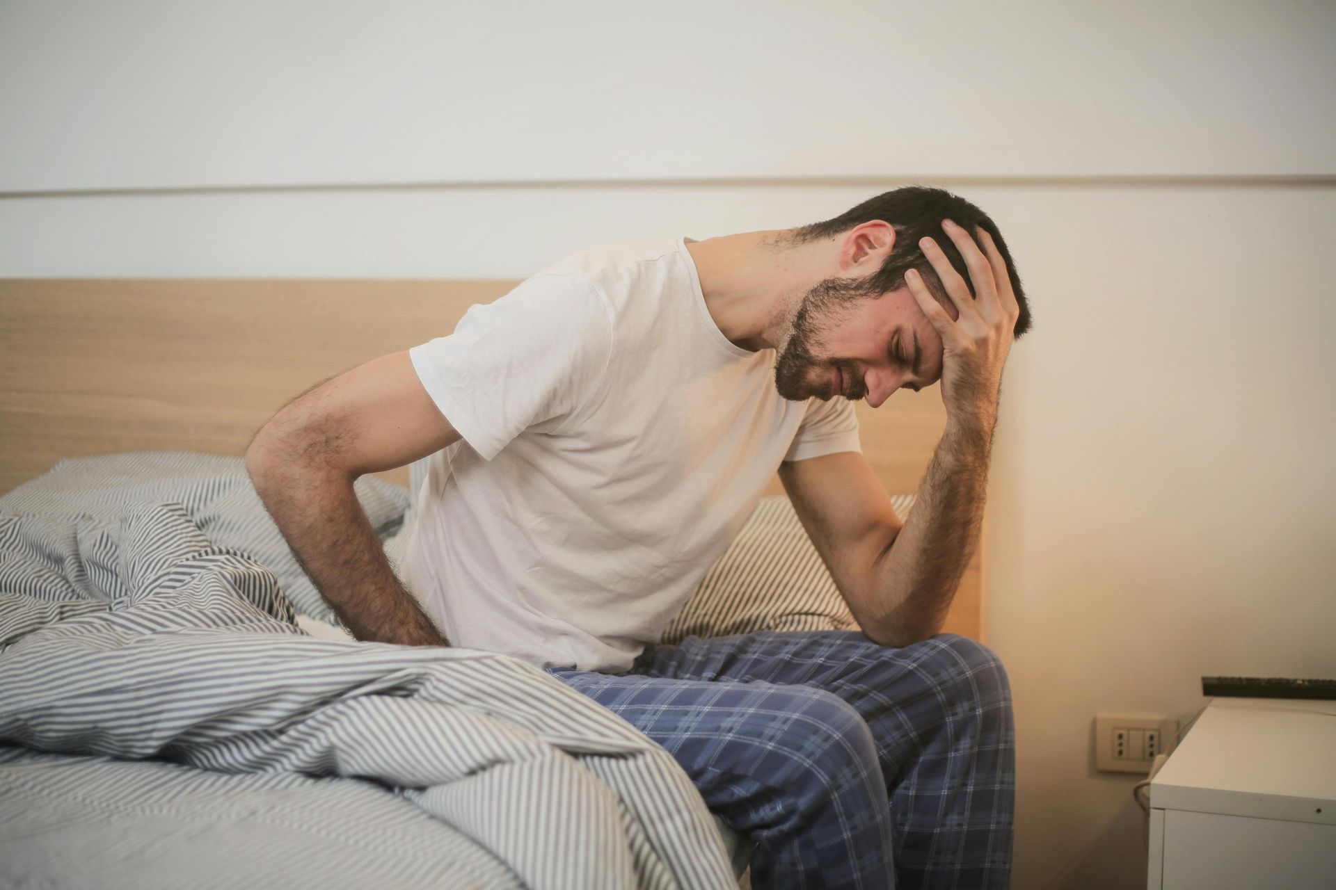 zieke man zittend op de rand van zijn bed in een blauw geruite pyjamabroek en een wit T-shirt met zi