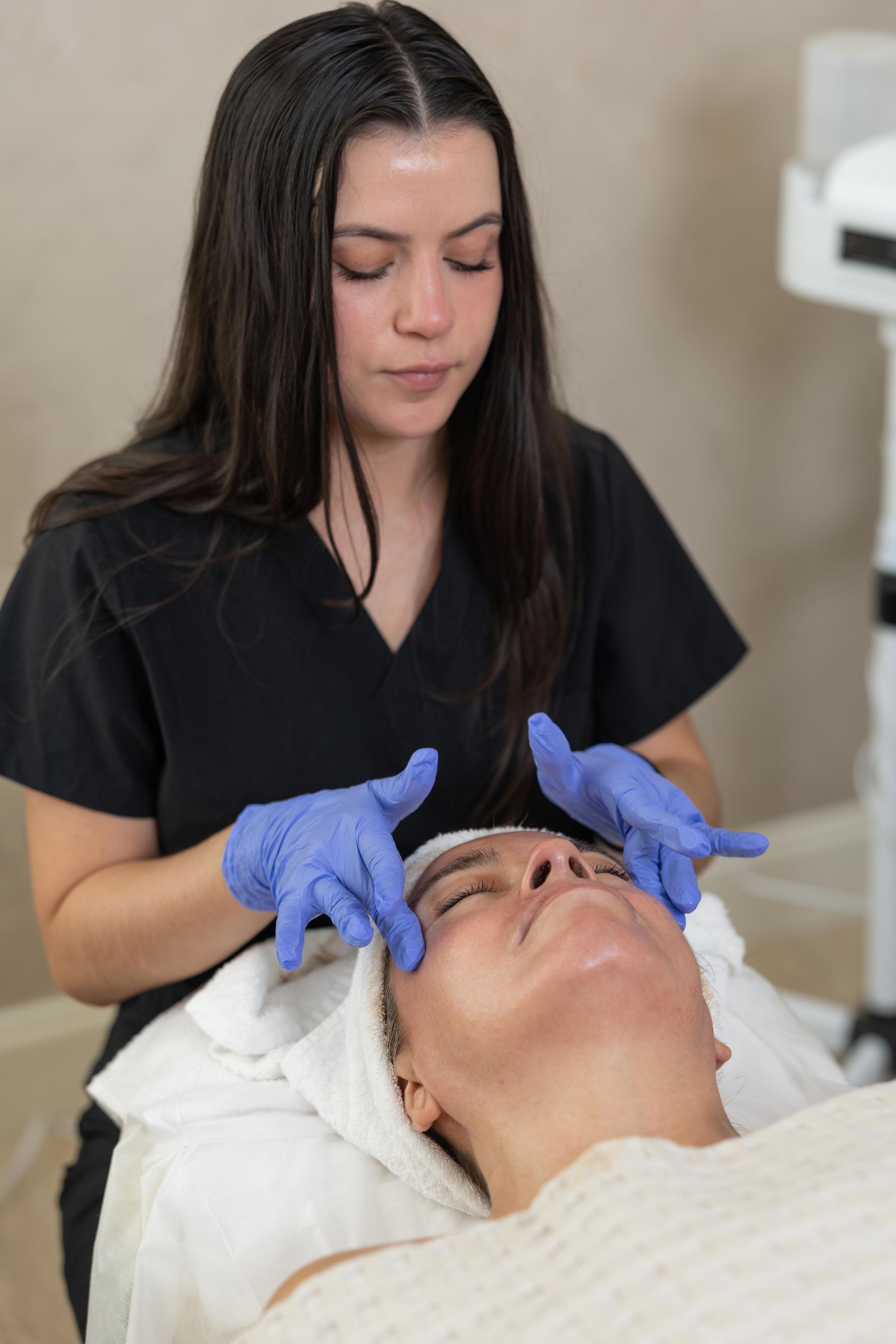 Aesthetician in black scrubs applying treatment to a clientβs face with gloved hands
