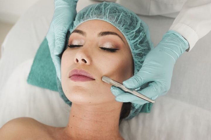 Cosmetic treatment on a woman’s face, with gloved hands applying a tool to her cheek while she lies back with eyes closed.
