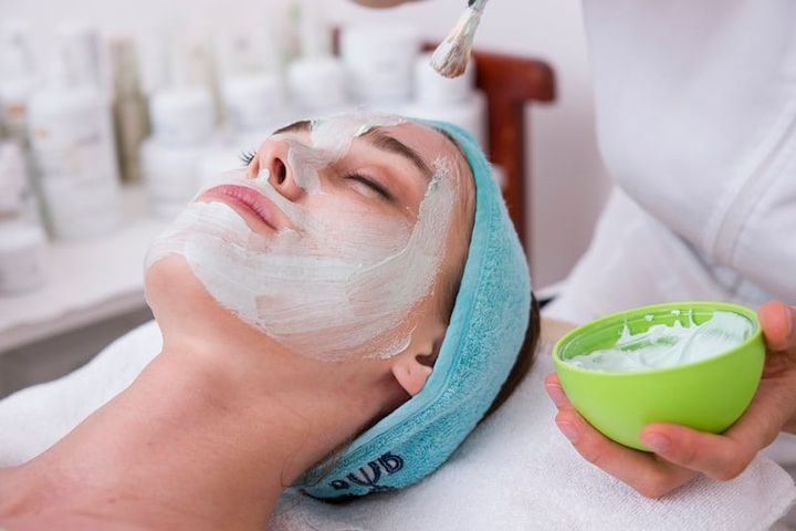 Woman receiving a facial mask treatment with a brush and green cream bowl in a spa