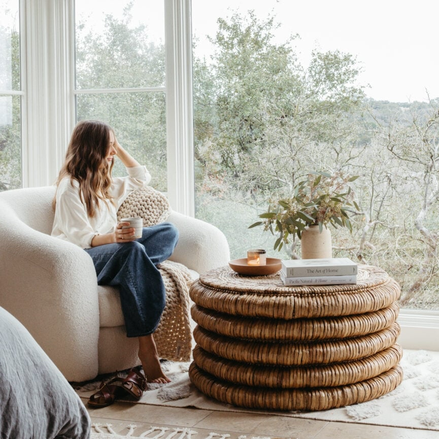 Woman sitting in a white armchair, looking out a large window at a tree-filled landscape. Brown coffee table in front.