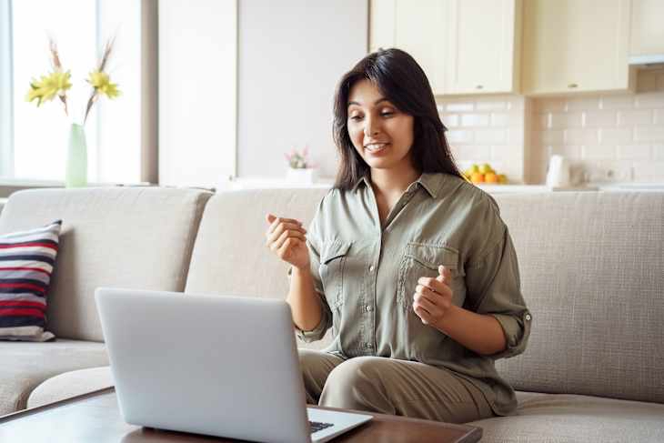 Woman in olive green shirt sits on couch, gesturing while looking at laptop.