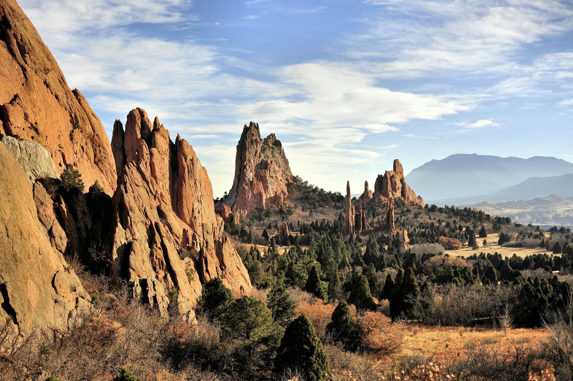 Red rock formations, trees, and distant mountains under a blue sky with clouds in Garden of the Gods, Colorado.