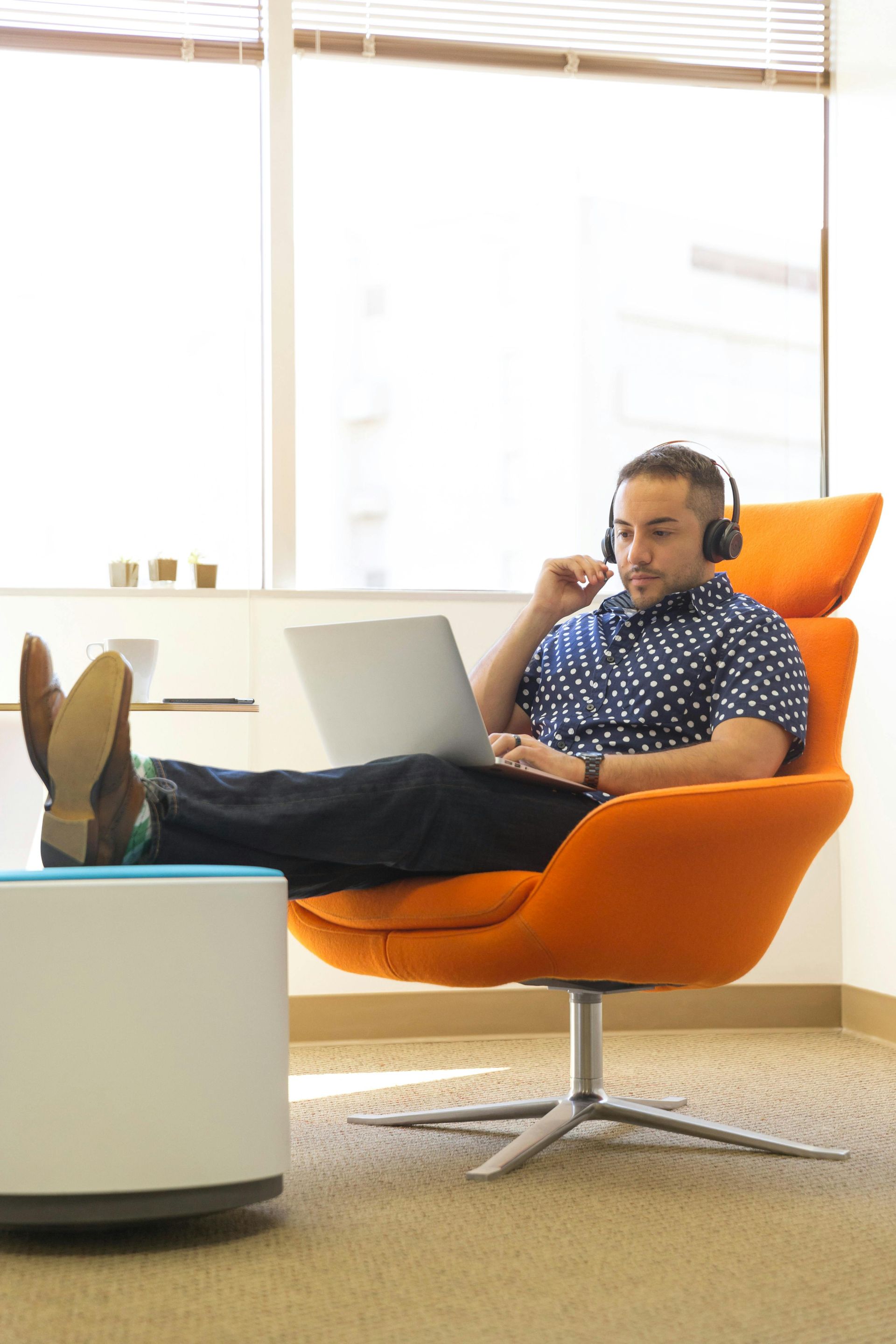Man wearing headphones relaxes in orange chair, feet on white drum, looking at laptop. Window background.