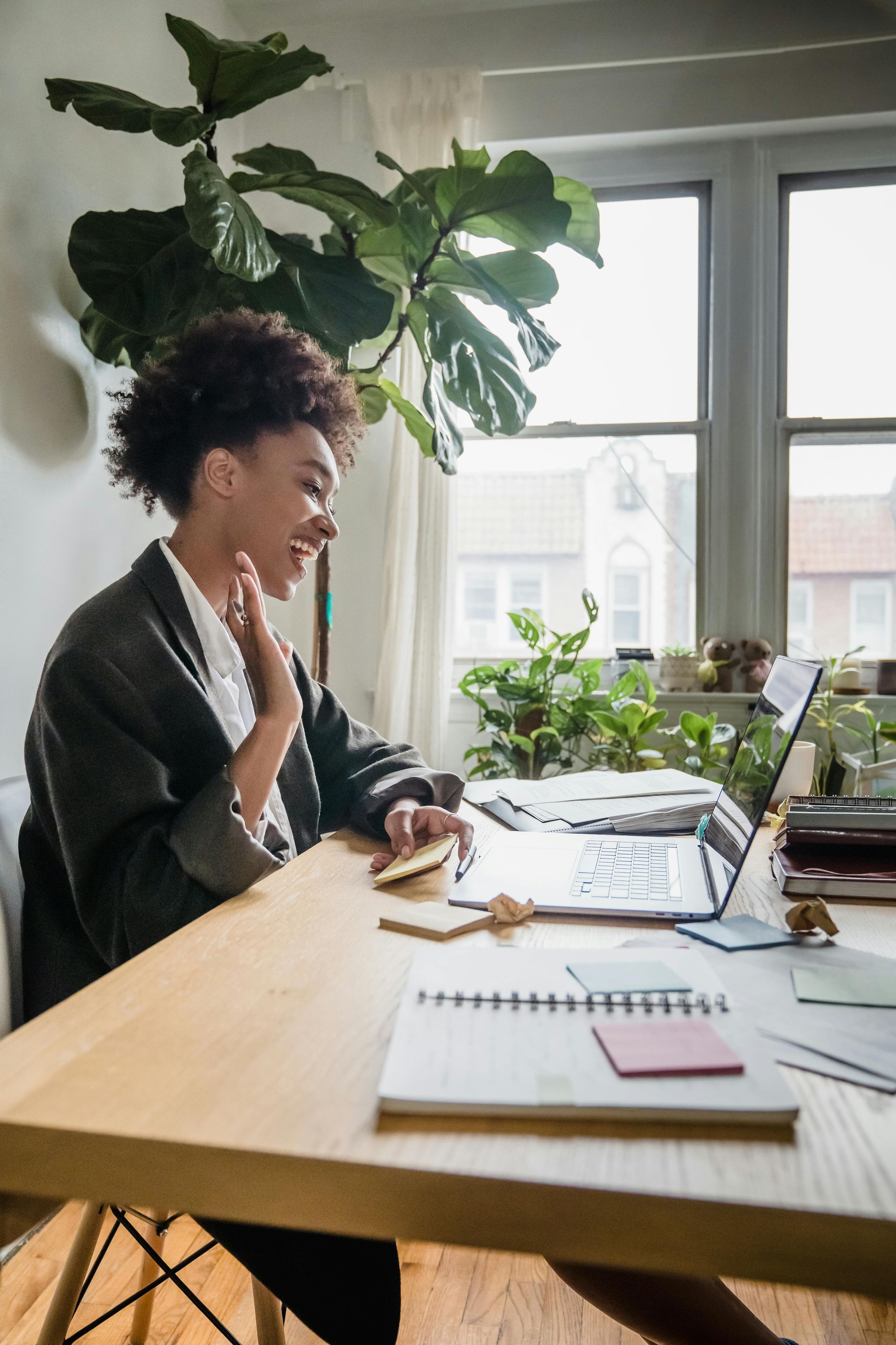 Woman waving during a video call on laptop at a desk with notebook and plants. Bright, indoor setting.