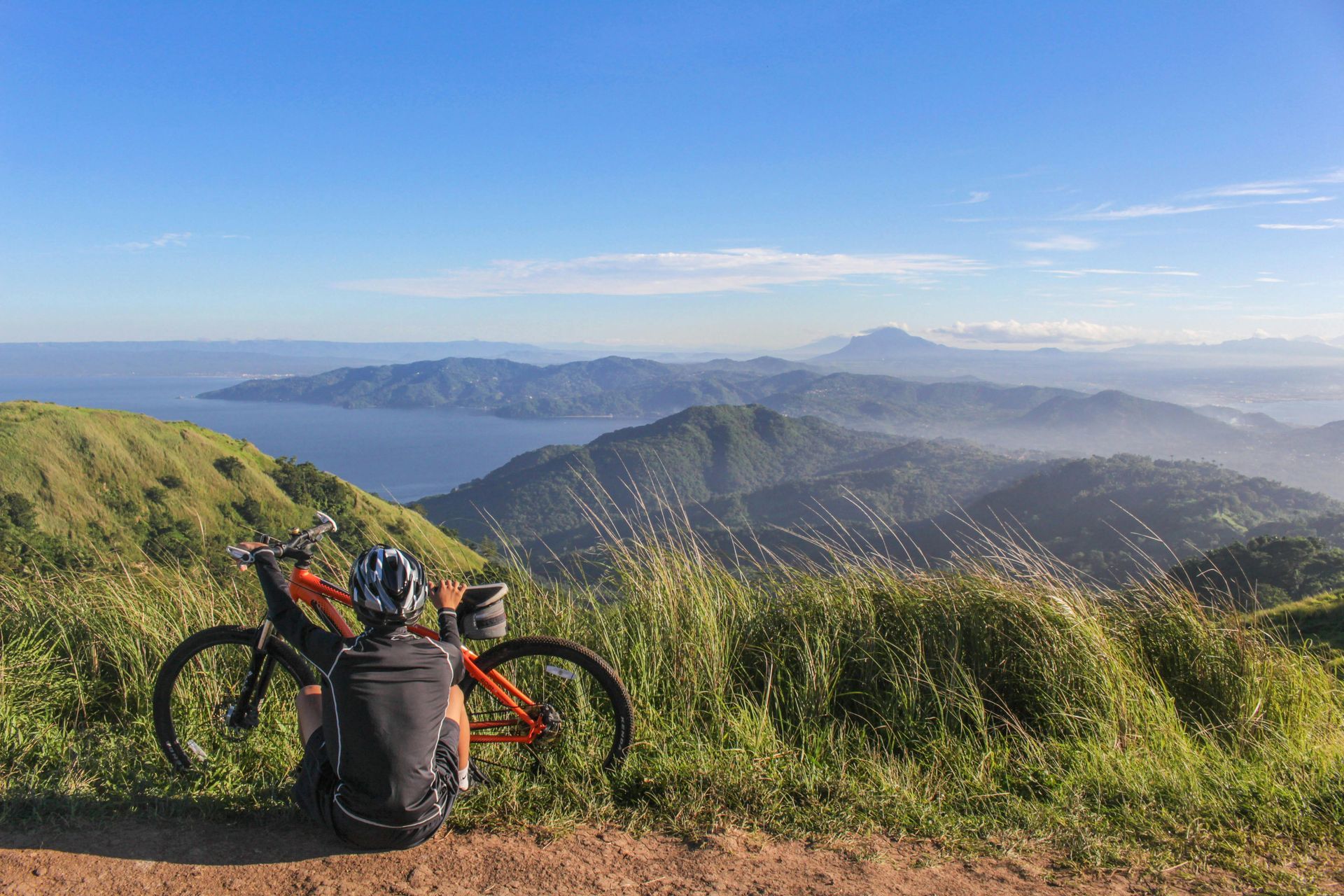 Person sits near an orange mountain bike on a hilltop, overlooking a scenic mountain and water vista under a blue sky.