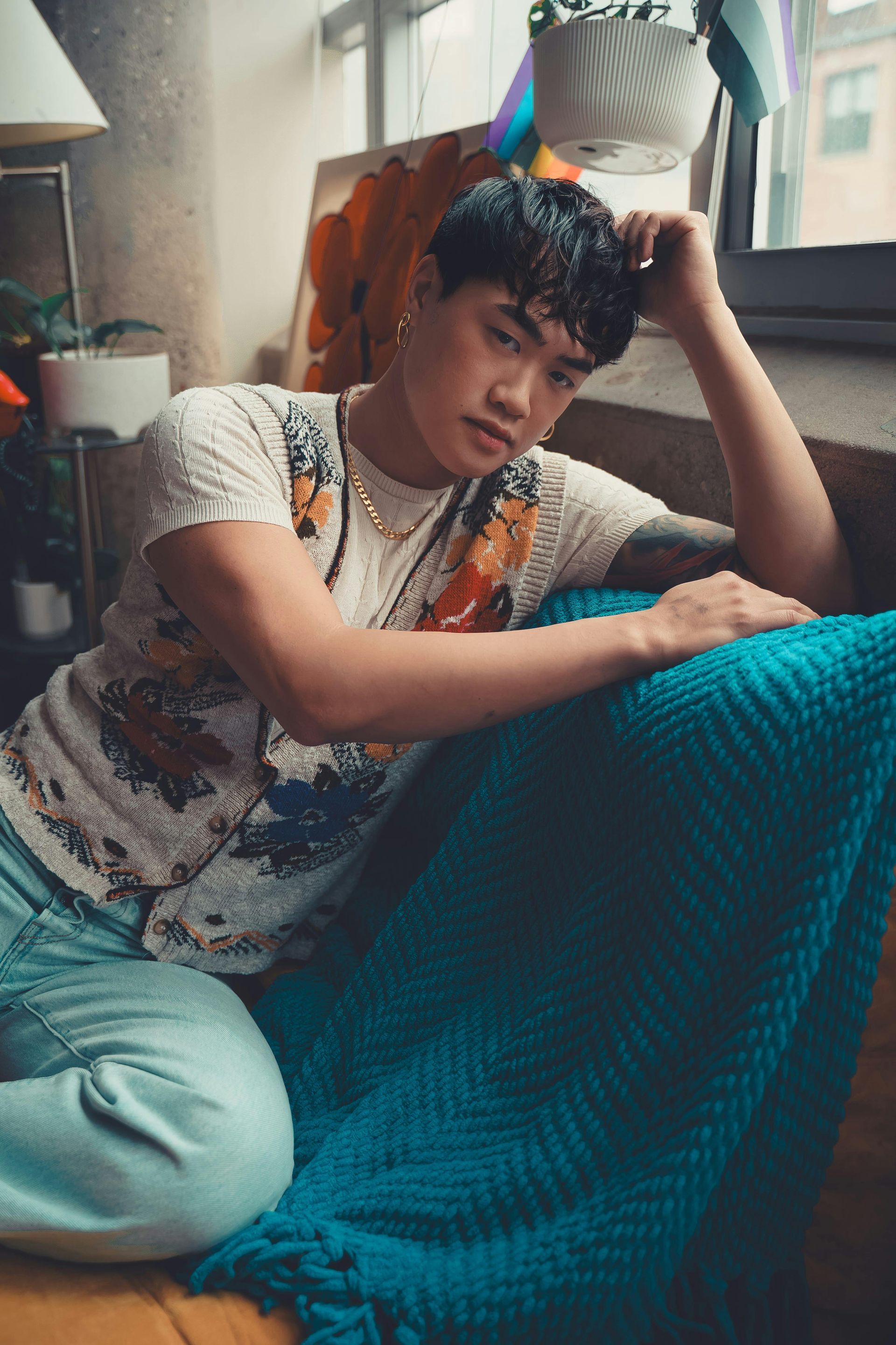 Person sitting on a blue couch, wearing a patterned shirt and jeans, leaning near a window.