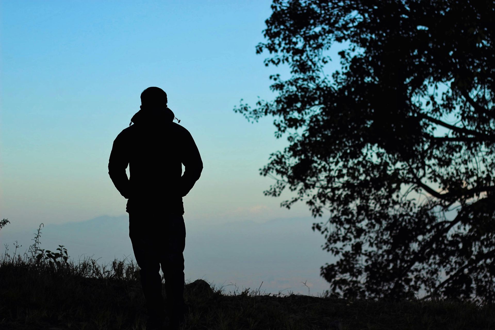 Silhouette of a person standing on a hill at dusk with a tree to the right and mountains in the background.