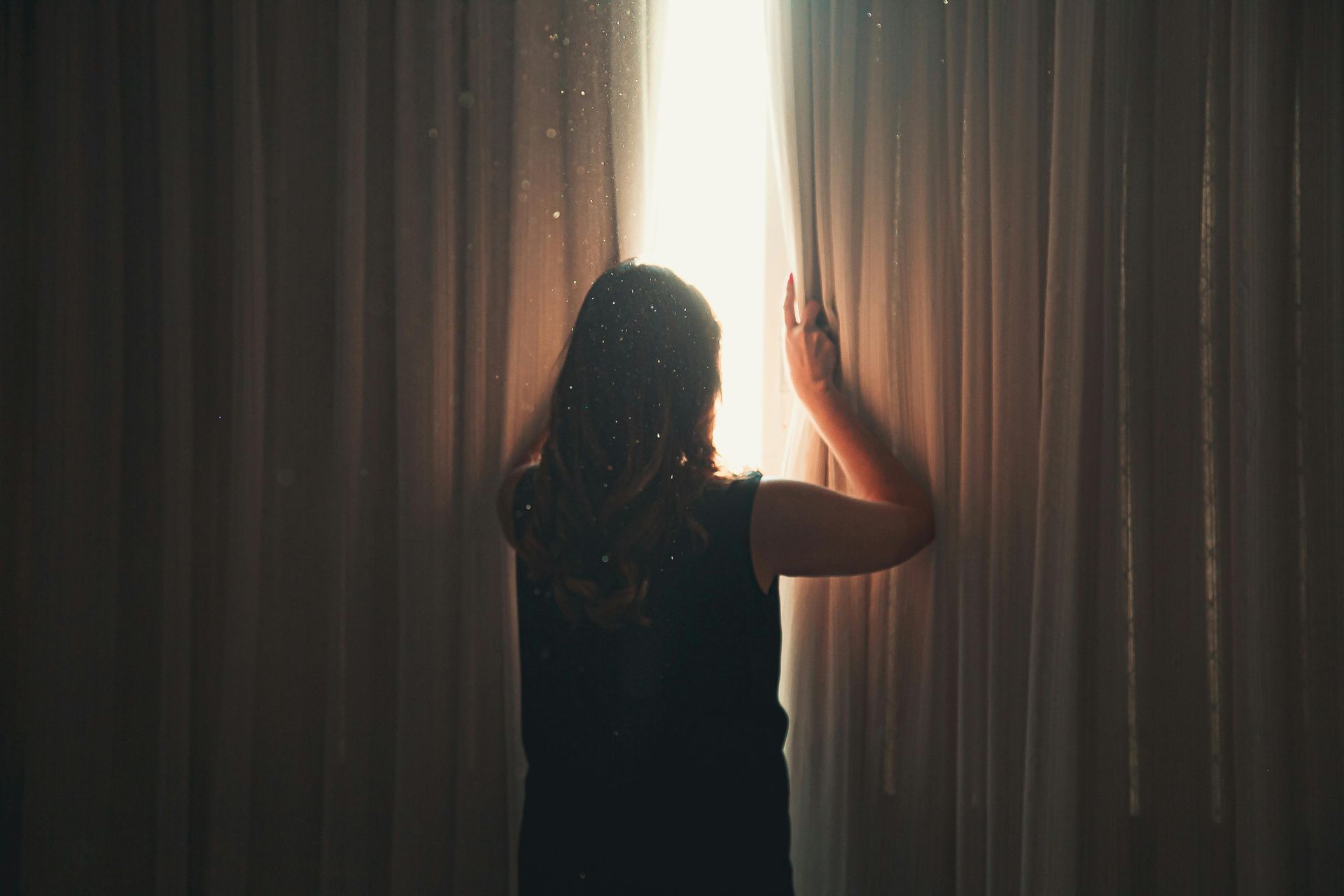 Woman stands before bright window, opening curtains. Sunlight streams through.