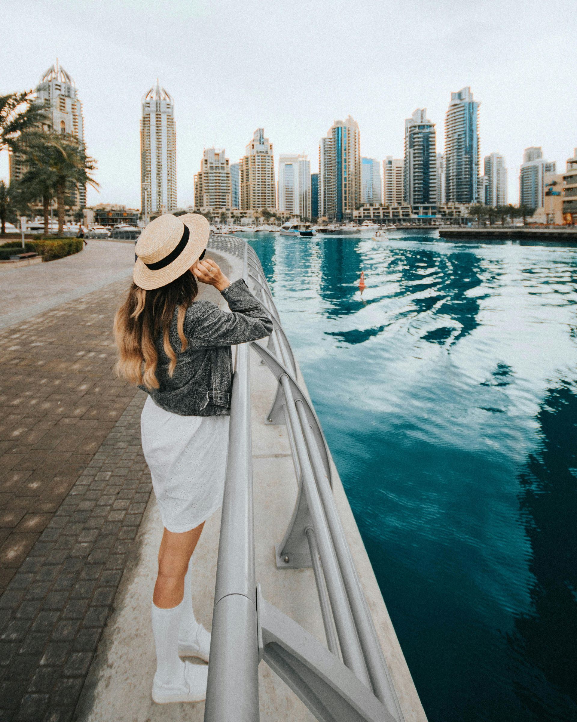 Woman in white dress and hat looks at cityscape over blue water.