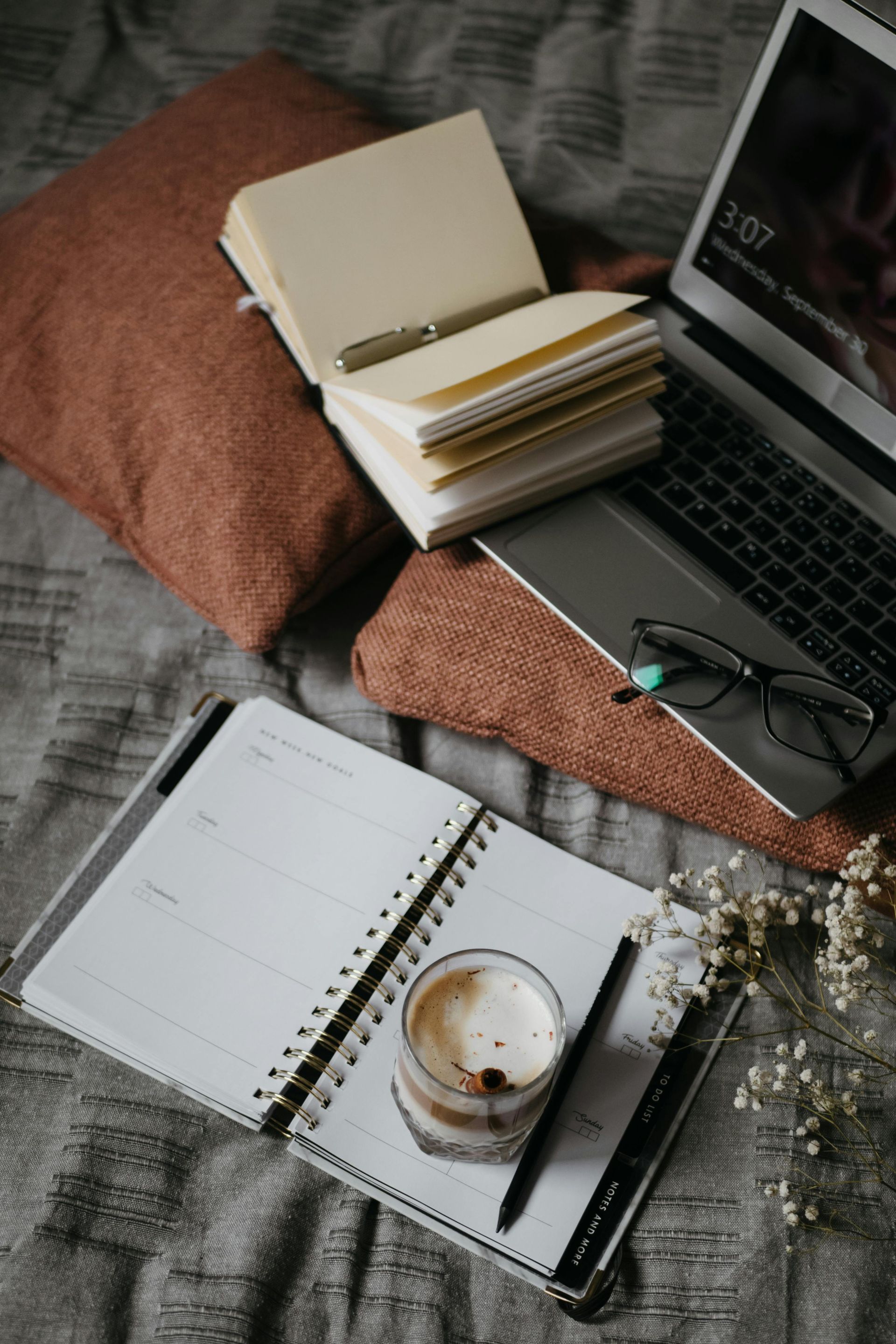 Laptop, notebooks, and coffee on a bed with pillows. Glasses and a pen are also present.